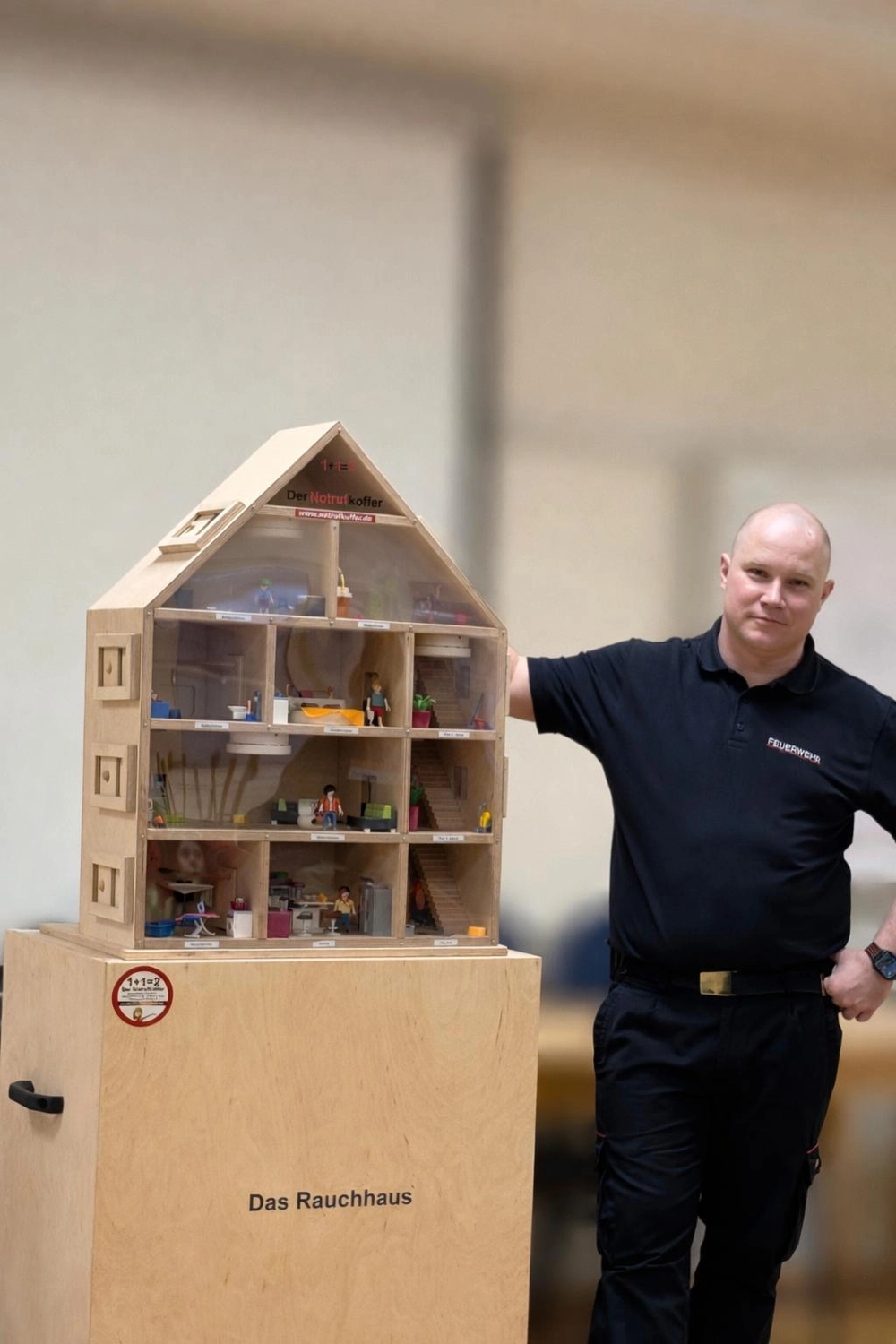Brandschutz Tobias Scheunemann - Brandschutz und Schulung - Ein Feuerwehrmann steht neben einem hölzernen Puppenhaus, das als Requisite für Brandverhaltensübungen dient.